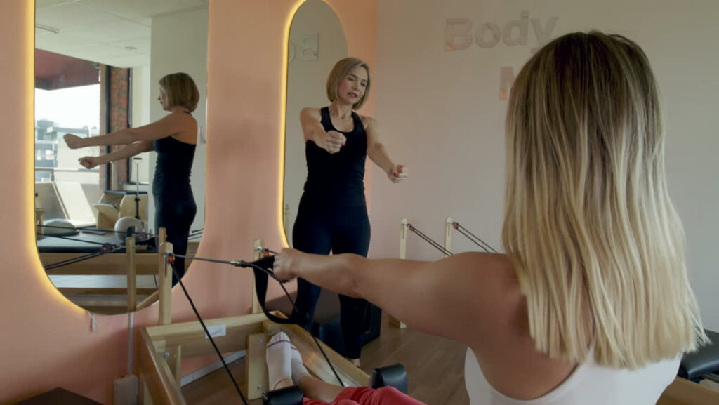 A patient works her shoulders on a reformer while the Physiotherapist demonstrates the arm position